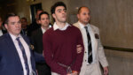 Young man in a maroon sweater is escorted down a hall.